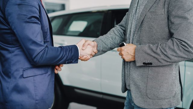 services-02 Two businessmen shaking hands in a car dealership, sealing a deal.